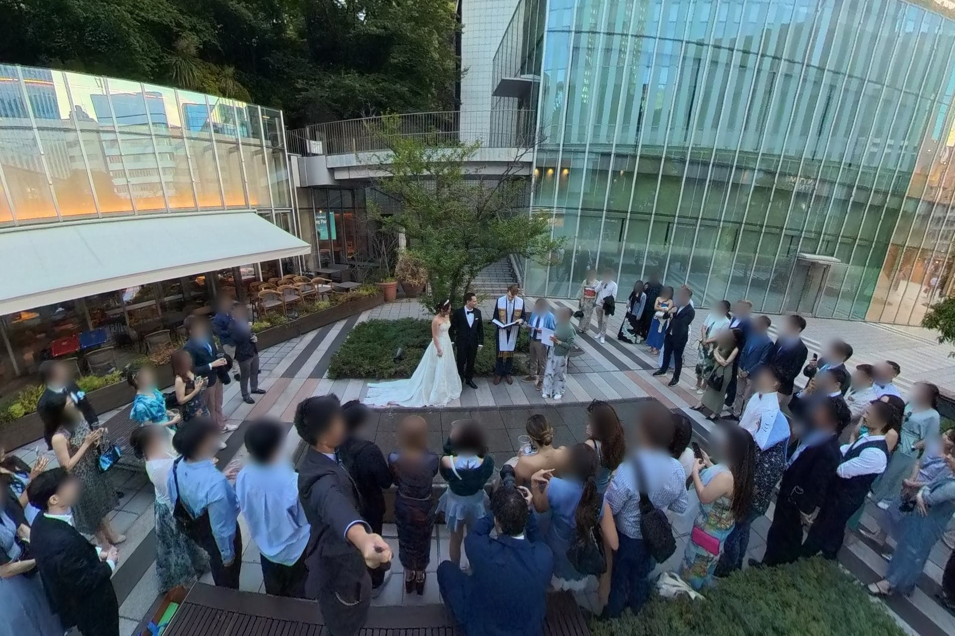 Vista ampia di una piccola cerimonia nuziale all'aperto in un cortile a Tokyo, con un amico che officia e gli invitati raccolti in cerchio intorno agli sposi