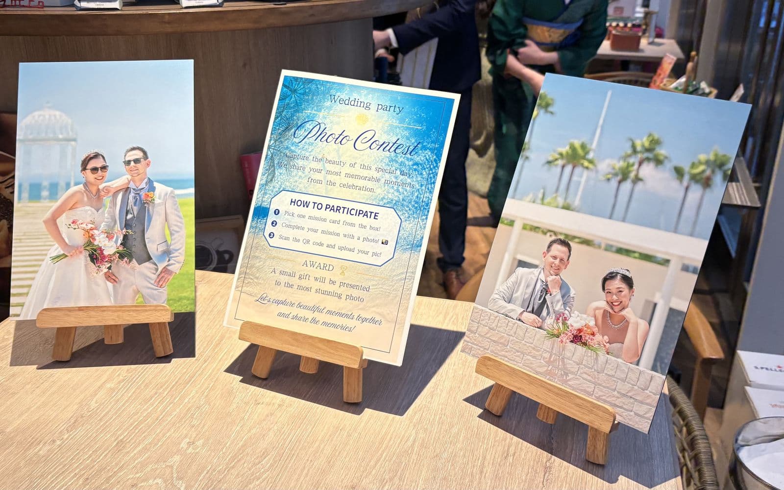Schild für den Fotowettbewerb beim Hochzeitsempfang, das erklärt, wie man eine Missionskarte zieht, das Foto macht und es mit einem QR-Code hochlädt, zwischen zwei gerahmten Fotos des Paares