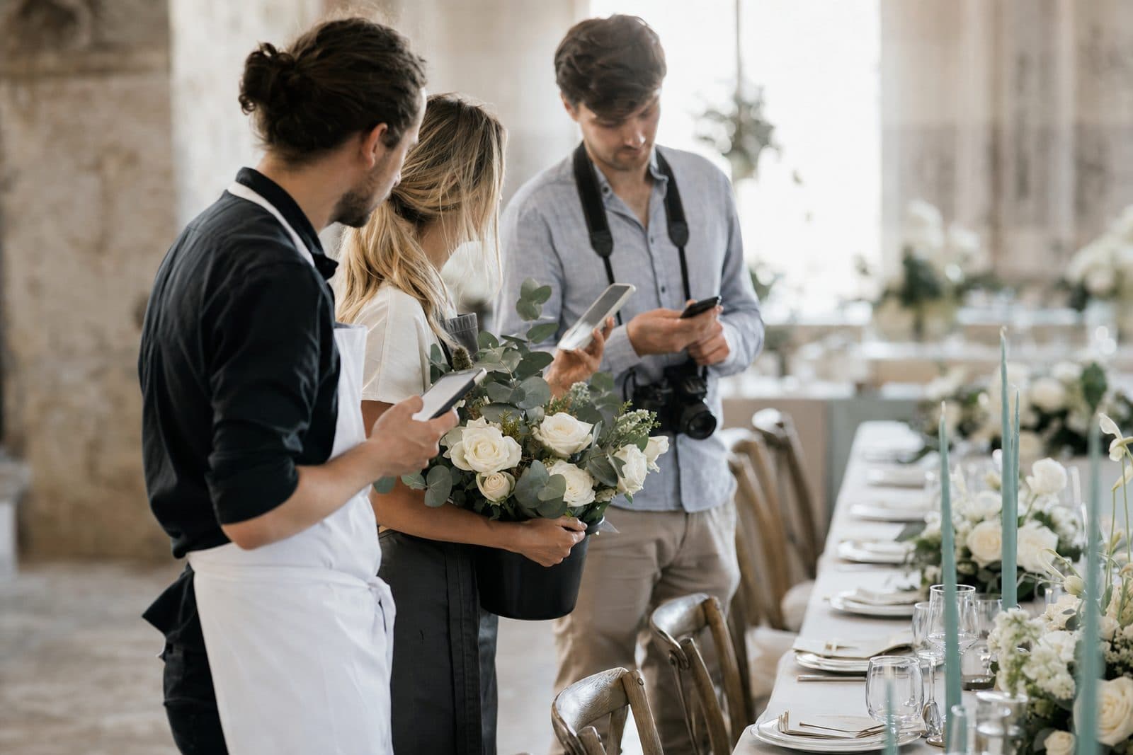 Un addetto al catering, una fiorista e un fotografo nella location del matrimonio, ciascuno intento a controllare il programma della giornata sul proprio telefono.